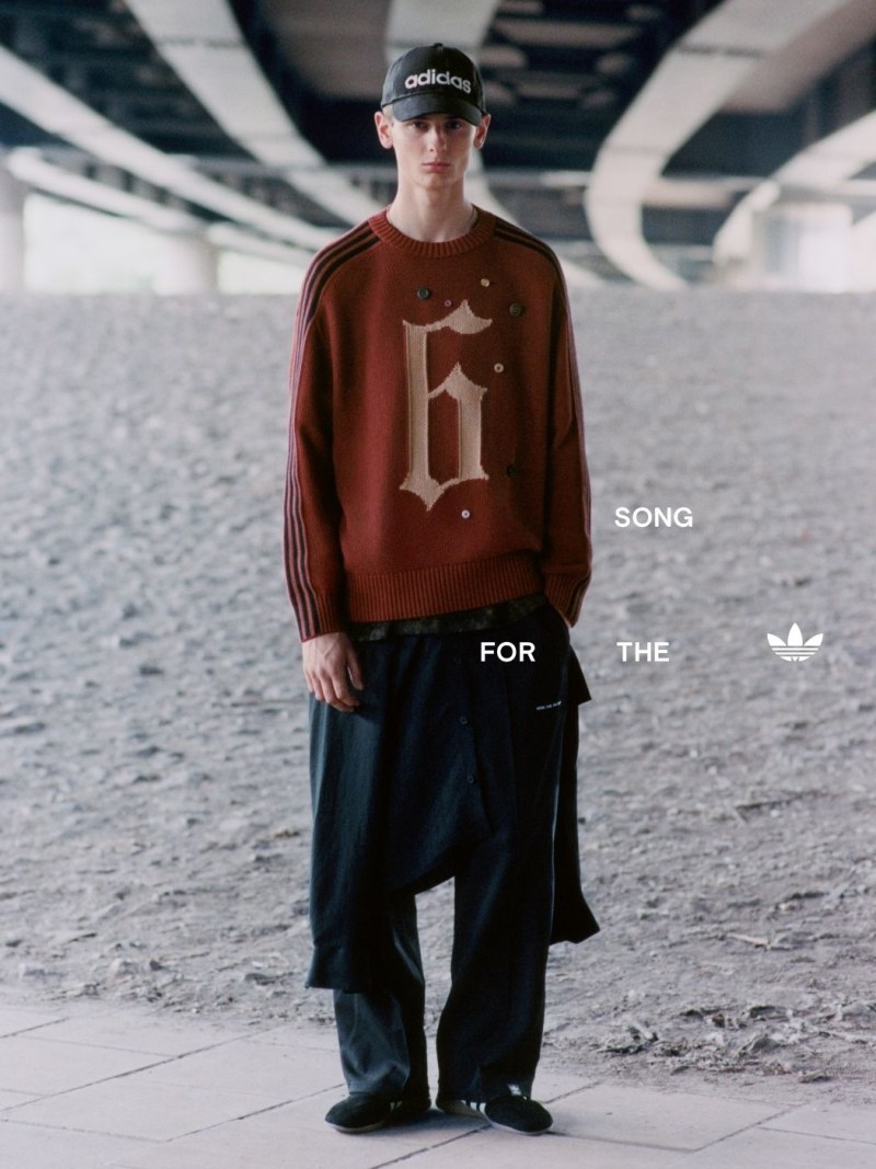 A person wearing a red sweater with a graphic design, an Adidas cap, and dark pants stands in a dimly lit indoor setting with a patterned floor.