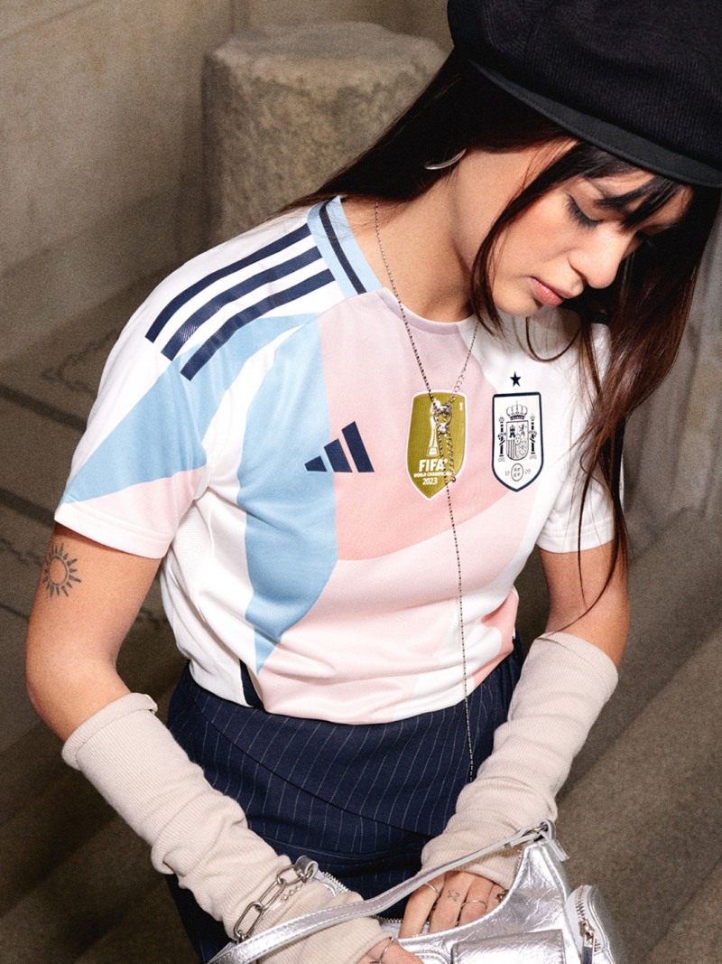 Na obrázku je mladá žena v růžovém a bílém sportovním dresu s logem adidas, která stojí před kamennou zdí.