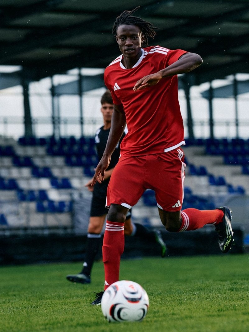 A soccer player in a red uniform is dribbling a ball on a soccer field with empty stadium seating visible in the background.