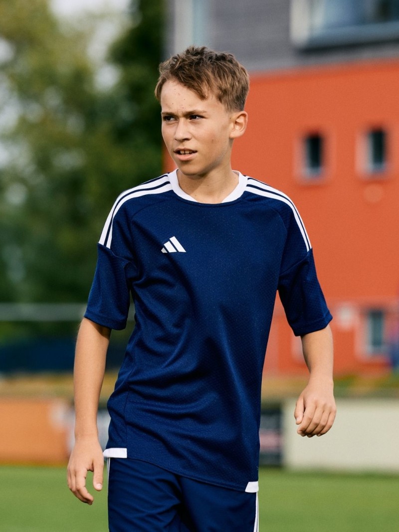 A young boy wearing a navy blue sports jersey stands on a grassy field in front of a colorful building.