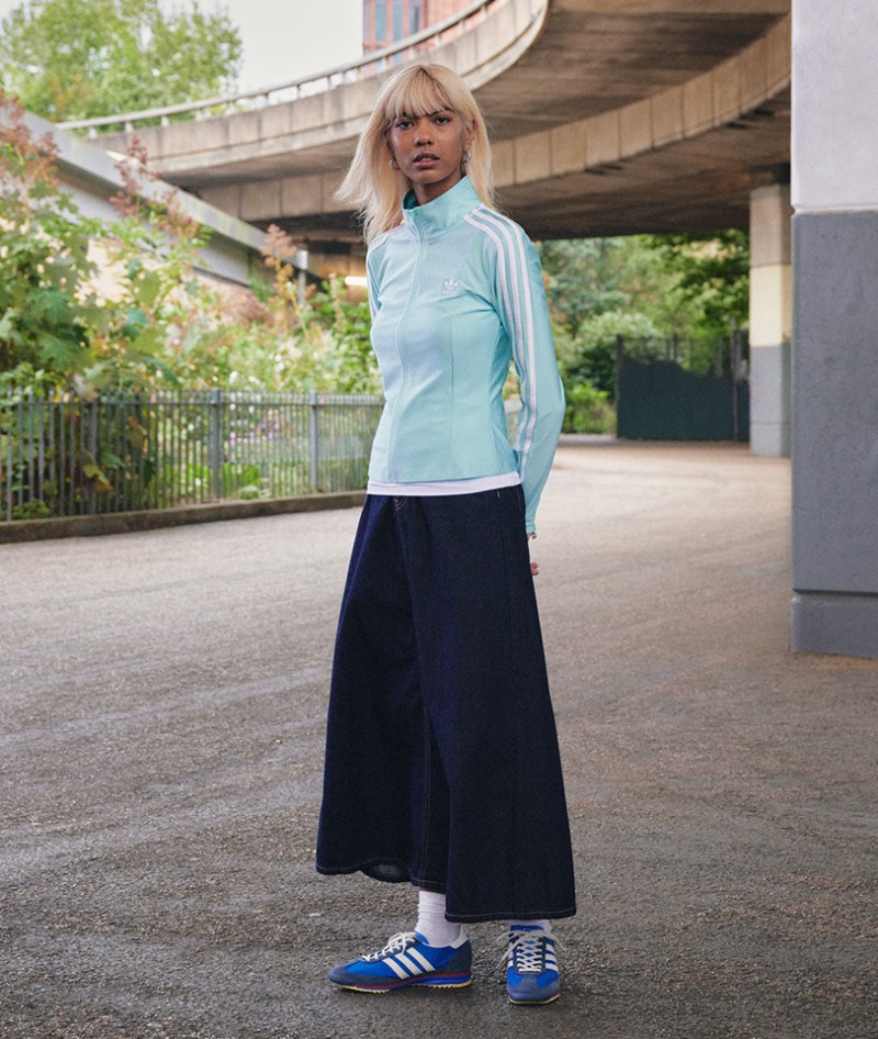 A young woman with blonde hair stands in front of a curved concrete structure, wearing a light blue top and a long navy blue skirt, paired with blue sneakers.