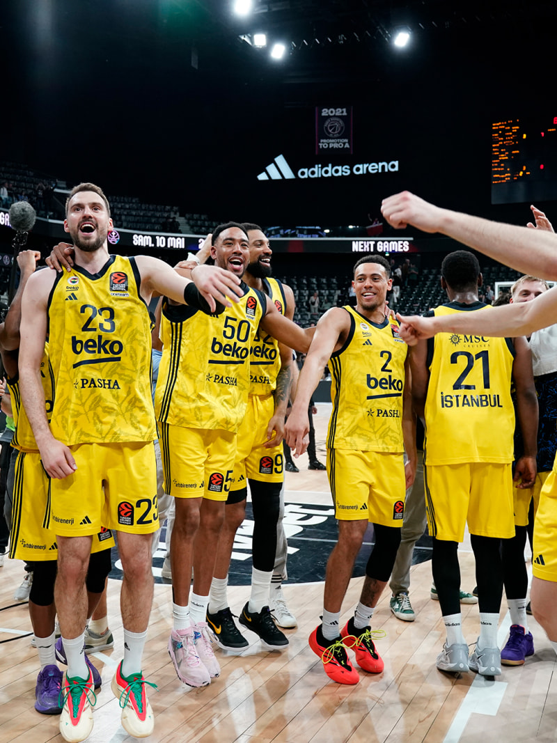 A group of basketball players in yellow uniforms celebrating on a basketball court with the Adidas arena logo visible in the background.