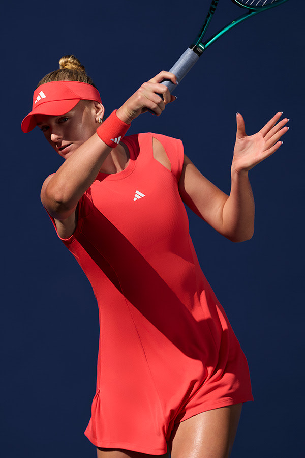 A female tennis player in a red outfit is swinging a tennis racket on a tennis court against a dark blue sky.
