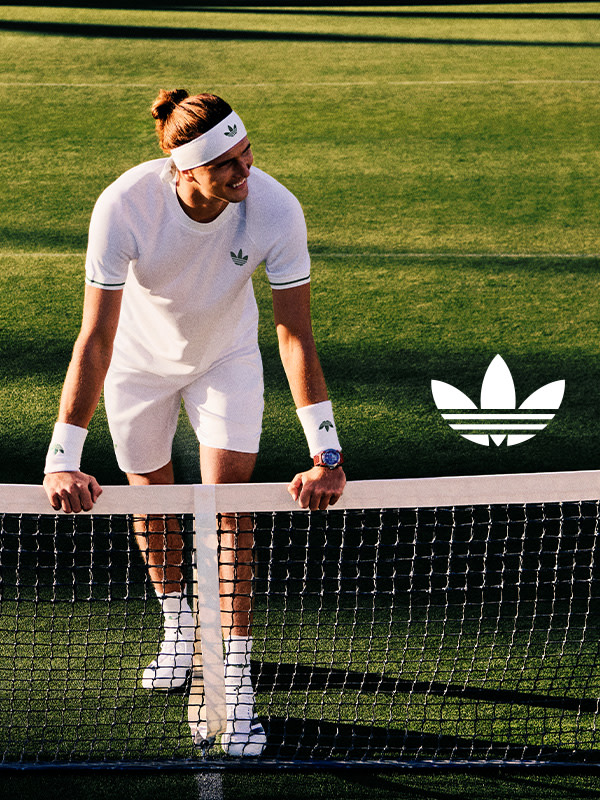 A person in white sportswear is standing on a tennis court, with the Adidas logo visible in the background.