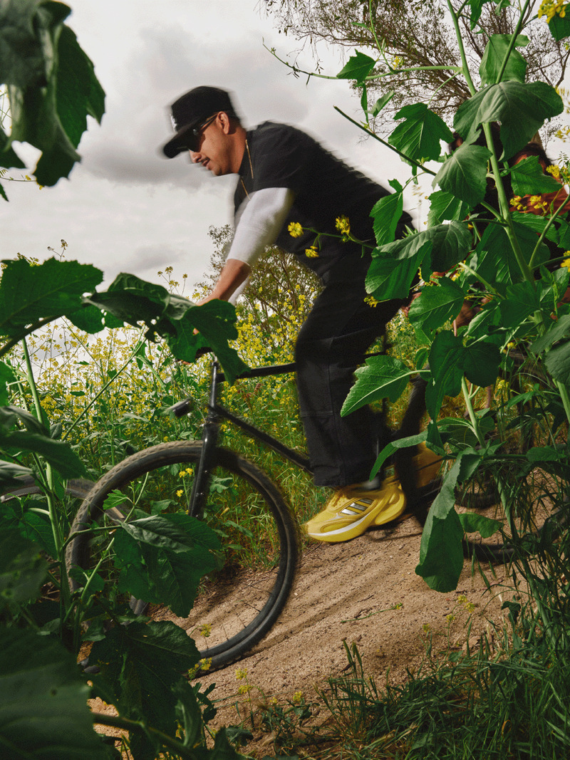 A person wearing black clothing and yellow shoes is riding a bicycle through a lush, overgrown garden environment with various plants and foliage in the background.