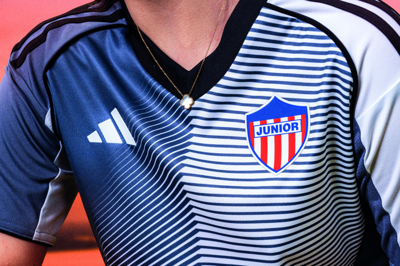 The image shows a blue and white striped jersey with the "Junior" logo prominently displayed on the front, set against a vibrant pink background.