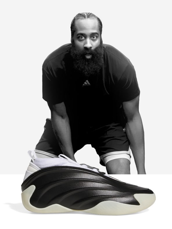 A bearded man in a black shirt sits on the floor, examining a large, curved sneaker in the foreground.