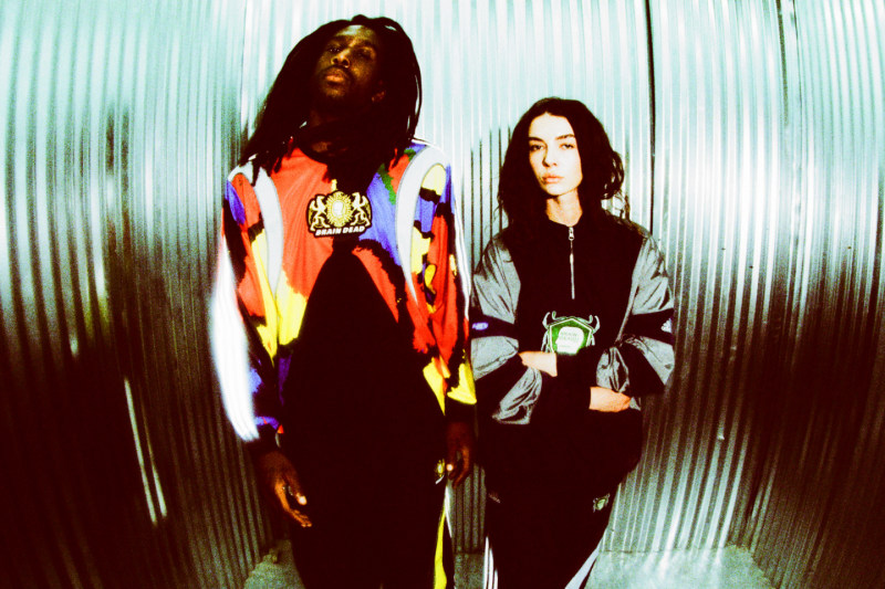 Two individuals, a person with long dark hair and another with shorter dark hair, stand in front of a blurred, reflective background.