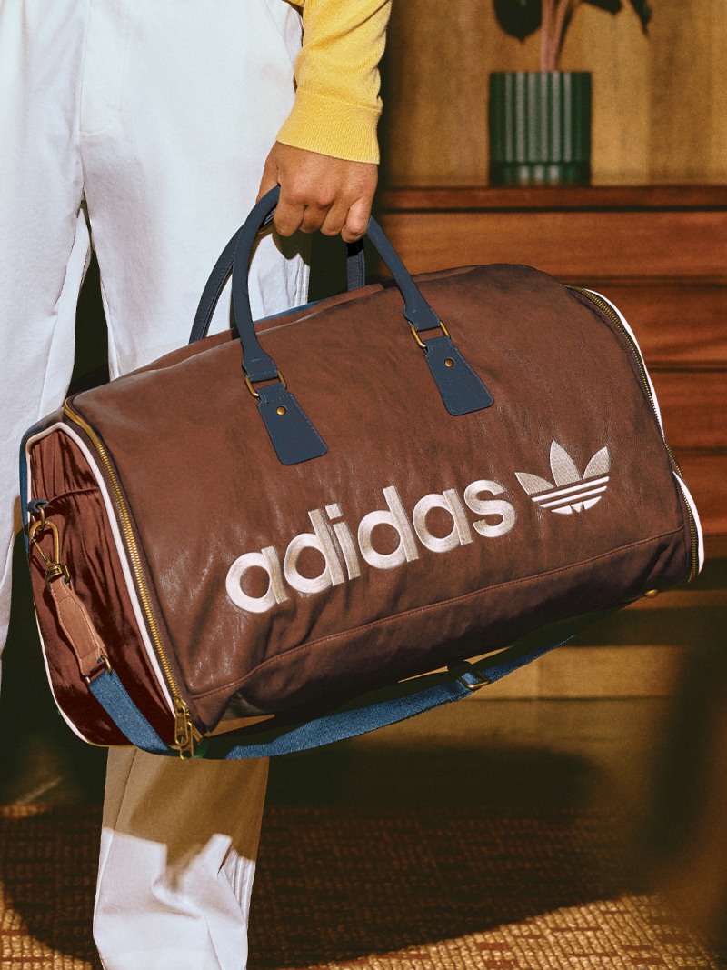 A person is holding a brown Adidas duffle bag in front of a wooden background.