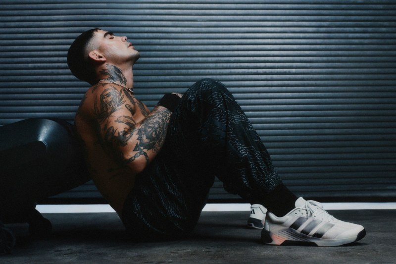 A person with tattoos sits on the ground in front of a metal shutter, wearing dark clothing and white sneakers.