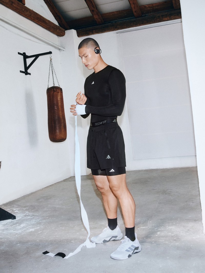A person in black athletic clothing stands in a room with wooden beams and a punching bag, appearing to be preparing for a workout.