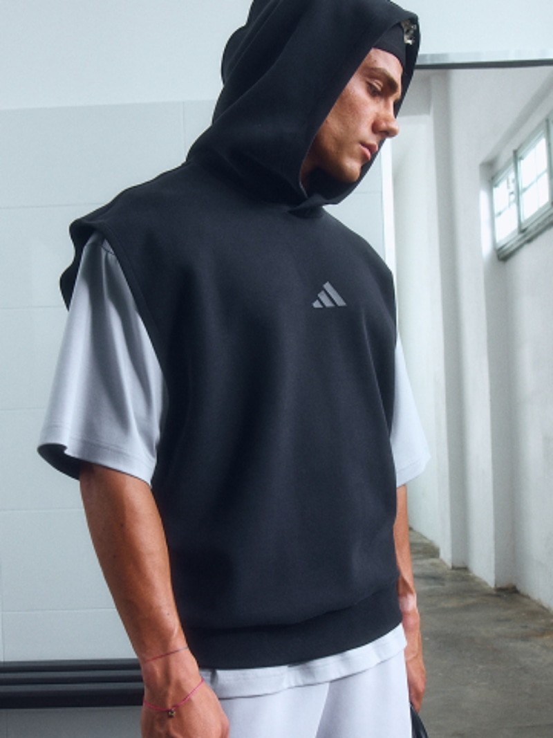 A person wearing a black hooded sweatshirt stands in a room with white walls and windows.