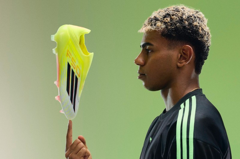 A young person with curly hair holding a bright yellow soccer cleat in front of a blurred green background.