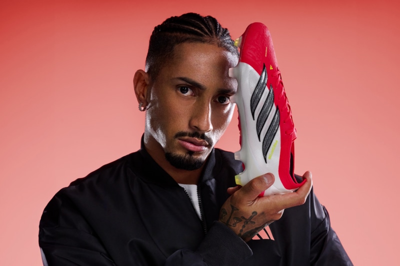 A man with facial tattoos and braided hair holds a large red and white striped candy cane against a vibrant red background.