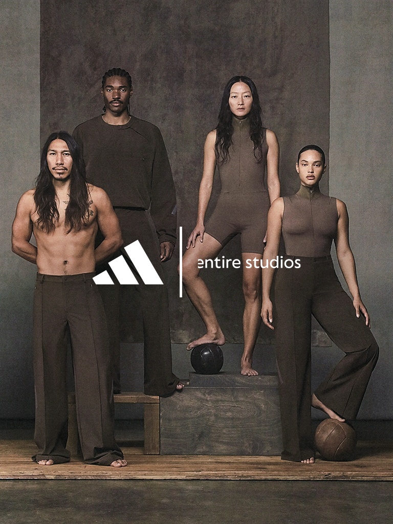 The image features a group of individuals, both male and female, posing together in a minimalist studio setting. The individuals are wearing dark-colored clothing, and the background is a plain, neutral-toned wall, creating a stark and dramatic visual contrast.