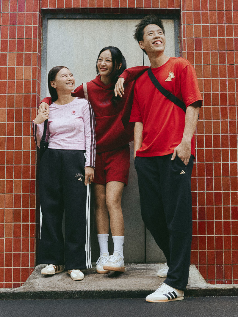 Three young people, two women and one man, are standing together in front of a red brick wall, all wearing red and black clothing and smiling happily.