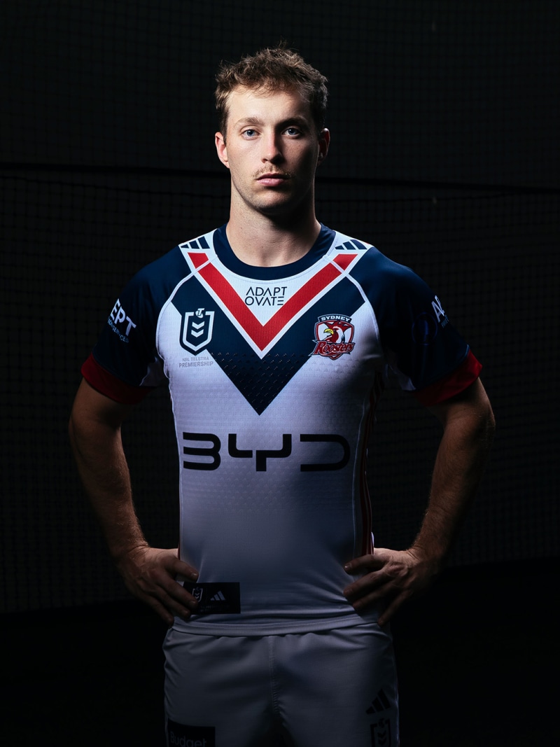 The image shows a young man wearing a sports jersey with the team's logo and sponsor prominently displayed, standing against a dark background.
