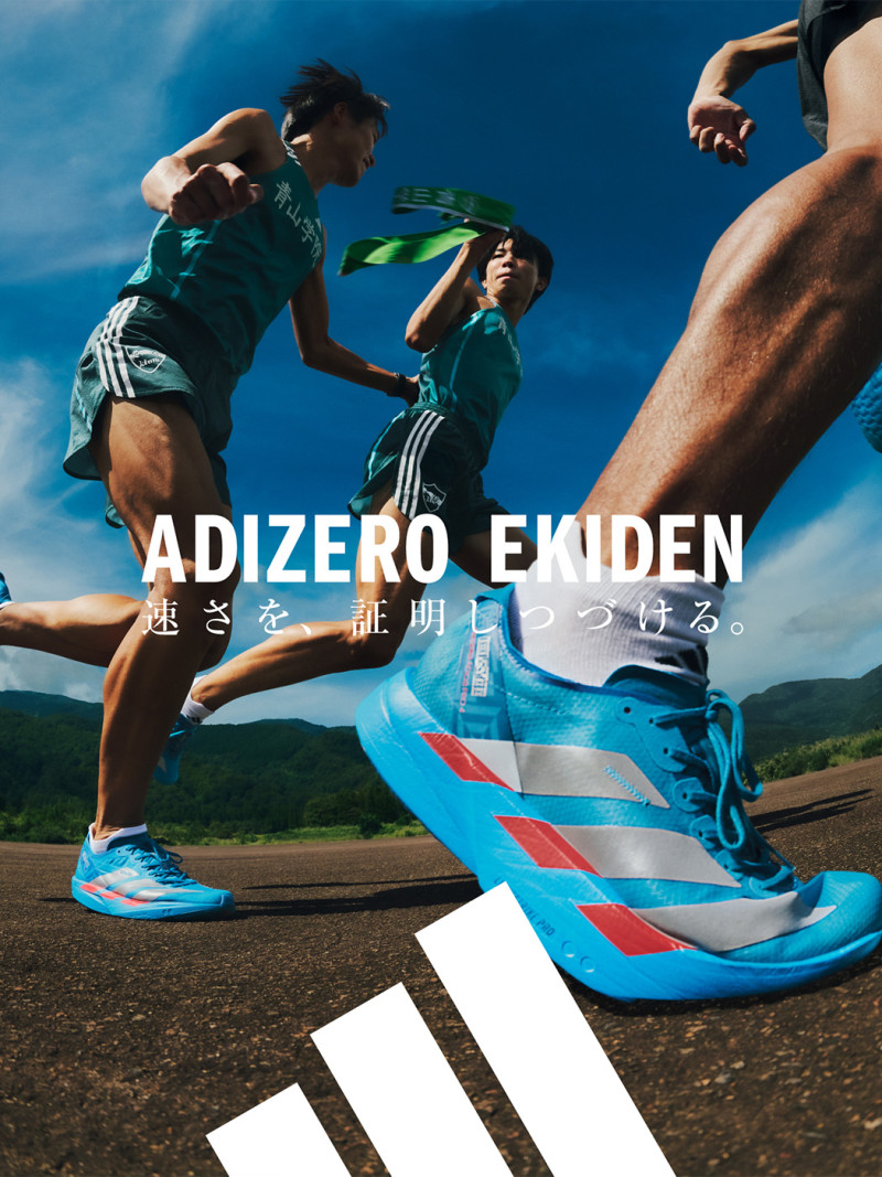 Two athletes in green uniforms are running on a road against a backdrop of a blue sky and mountainous terrain.