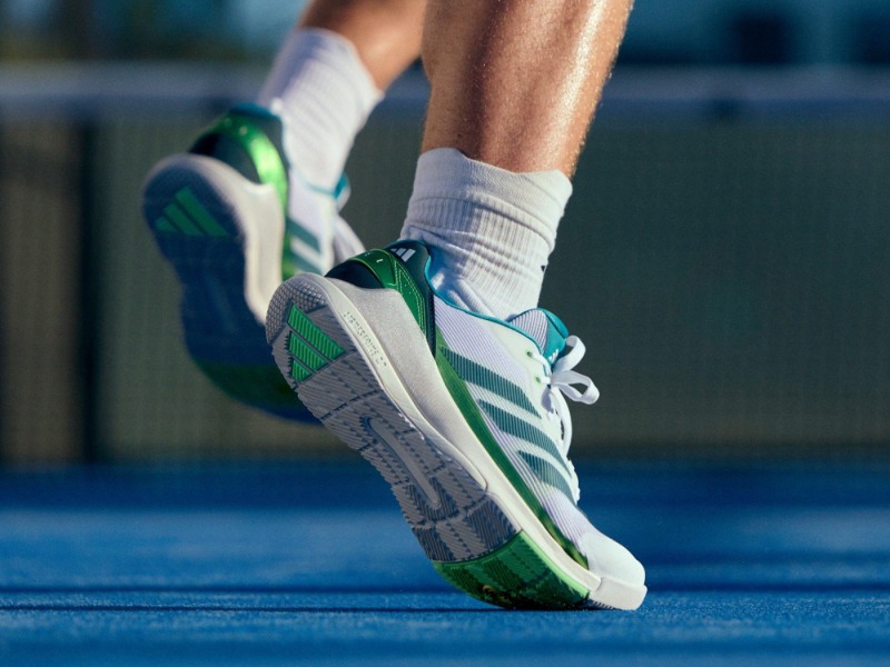 A pair of white and green athletic shoes with a blue and gray sole are visible on a blue surface, likely a sports court or track.