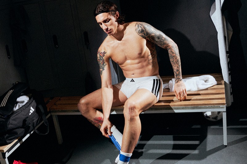A shirtless, tattooed individual with a muscular physique sits on a bench in a dimly lit room, wearing white shorts and blue socks.