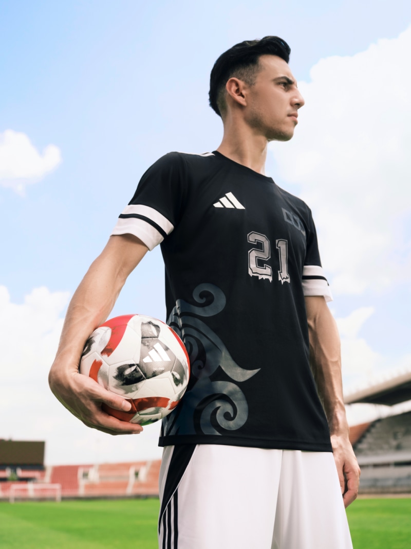 A young man in a black sports jersey holding a soccer ball on a sports field with a cloudy sky in the background.