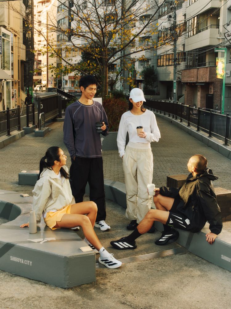 A group of people, some sitting and some standing, in a city street setting with buildings and trees visible in the background.