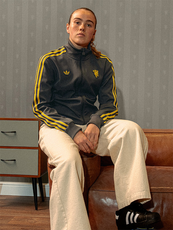 A person wearing a black and yellow tracksuit jacket is seated on a wooden bench against a plain gray background.