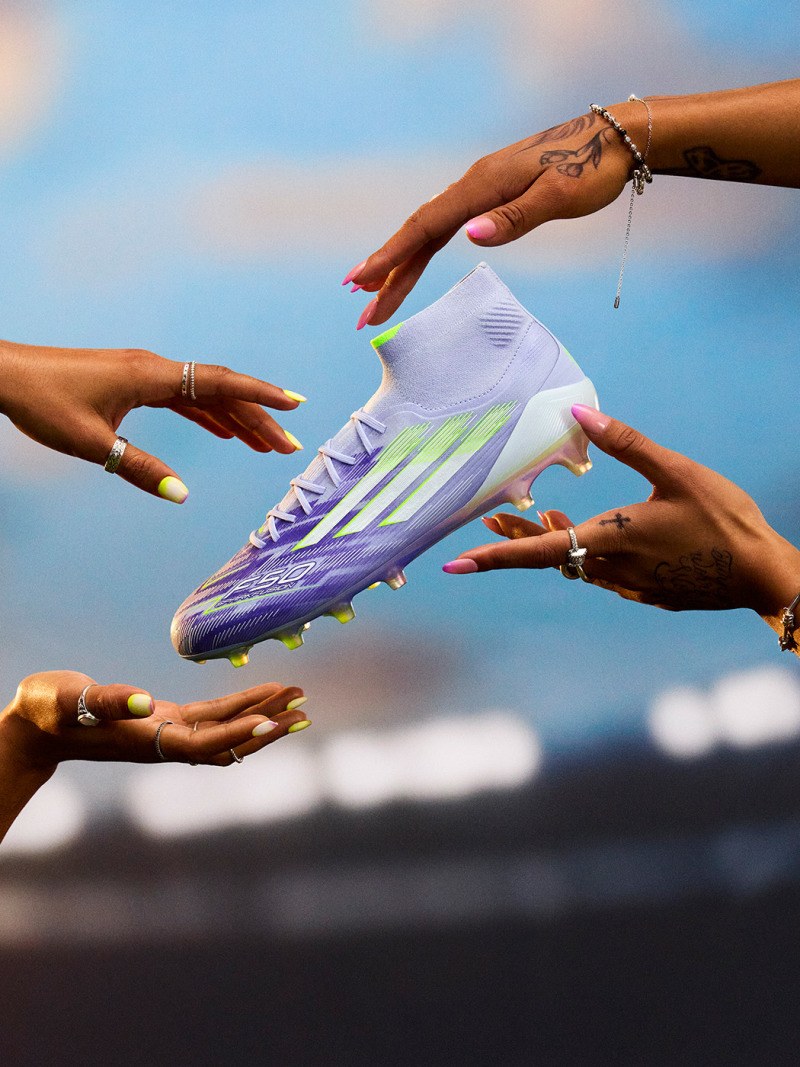 The image depicts a soccer cleat being passed between multiple hands against a backdrop of a cloudy sky.