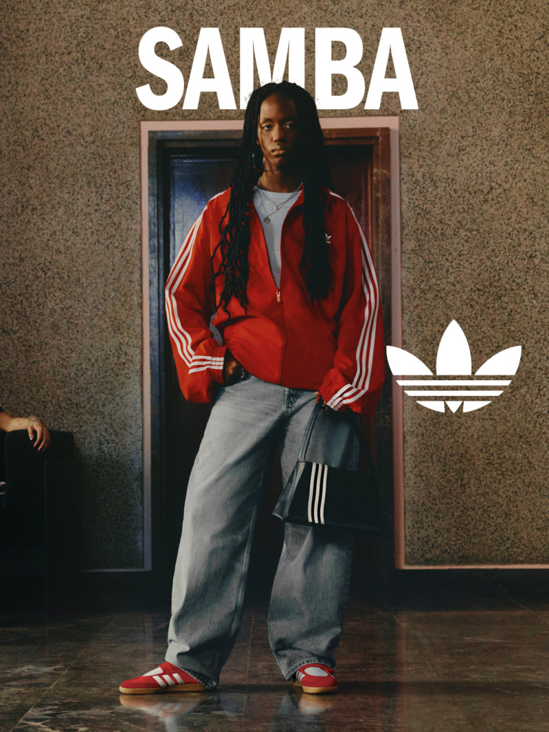 A person wearing a red Adidas jacket and gray sweatpants stands in front of a wall with the word "SAMBA" prominently displayed.