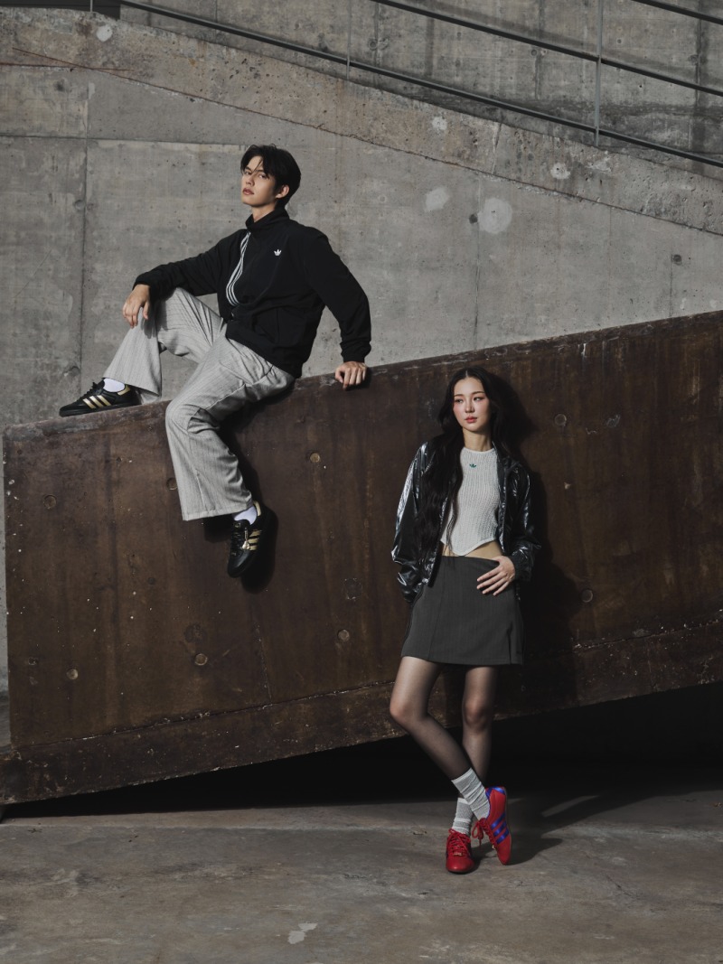 Two individuals, a man and a woman, are posing in an urban setting with a concrete wall in the background.