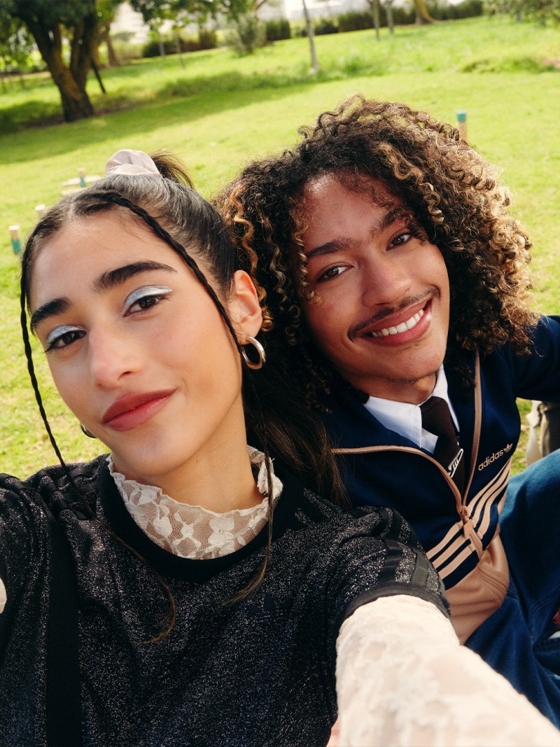 Two young women with warm, friendly expressions are smiling and posing together in a grassy, tree-lined outdoor setting.