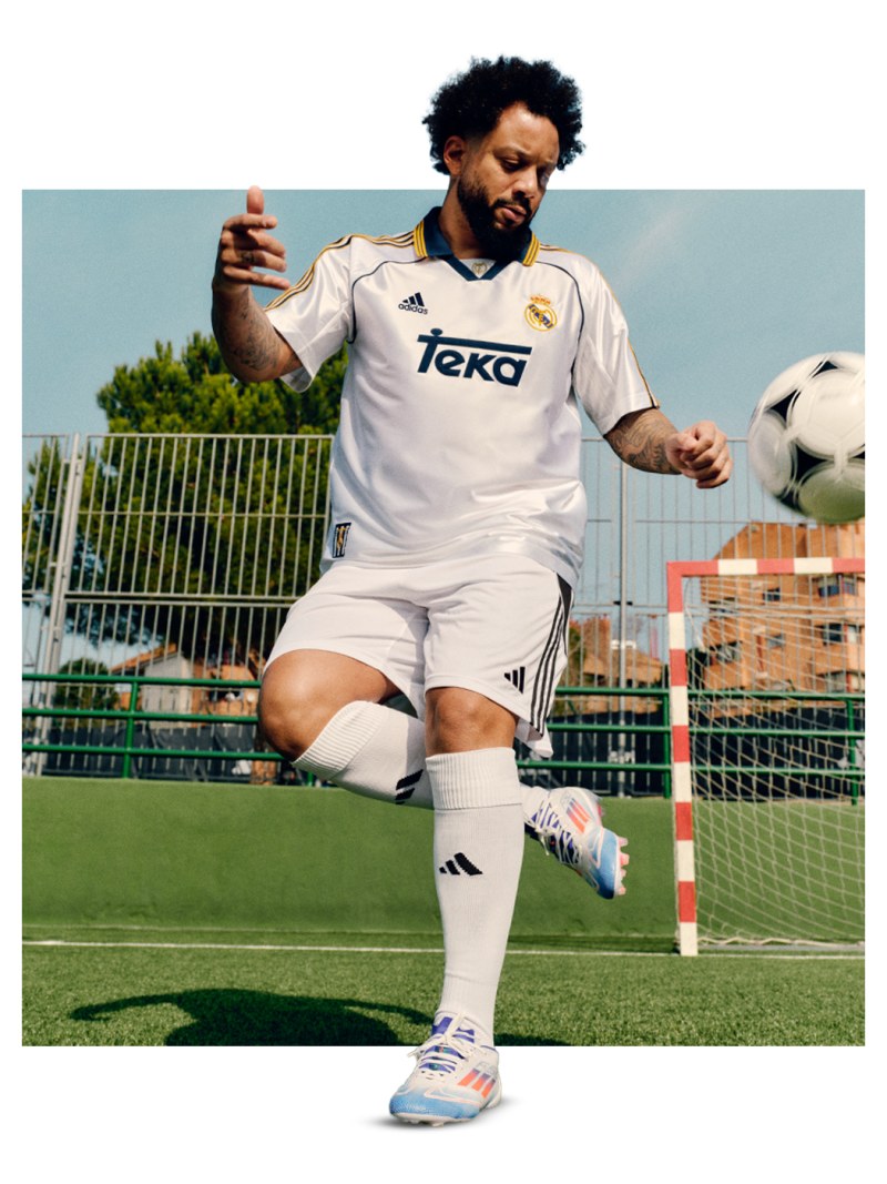 A soccer player in a white jersey and shorts is standing on a soccer field, with a soccer ball at his feet and a goal post visible in the background.