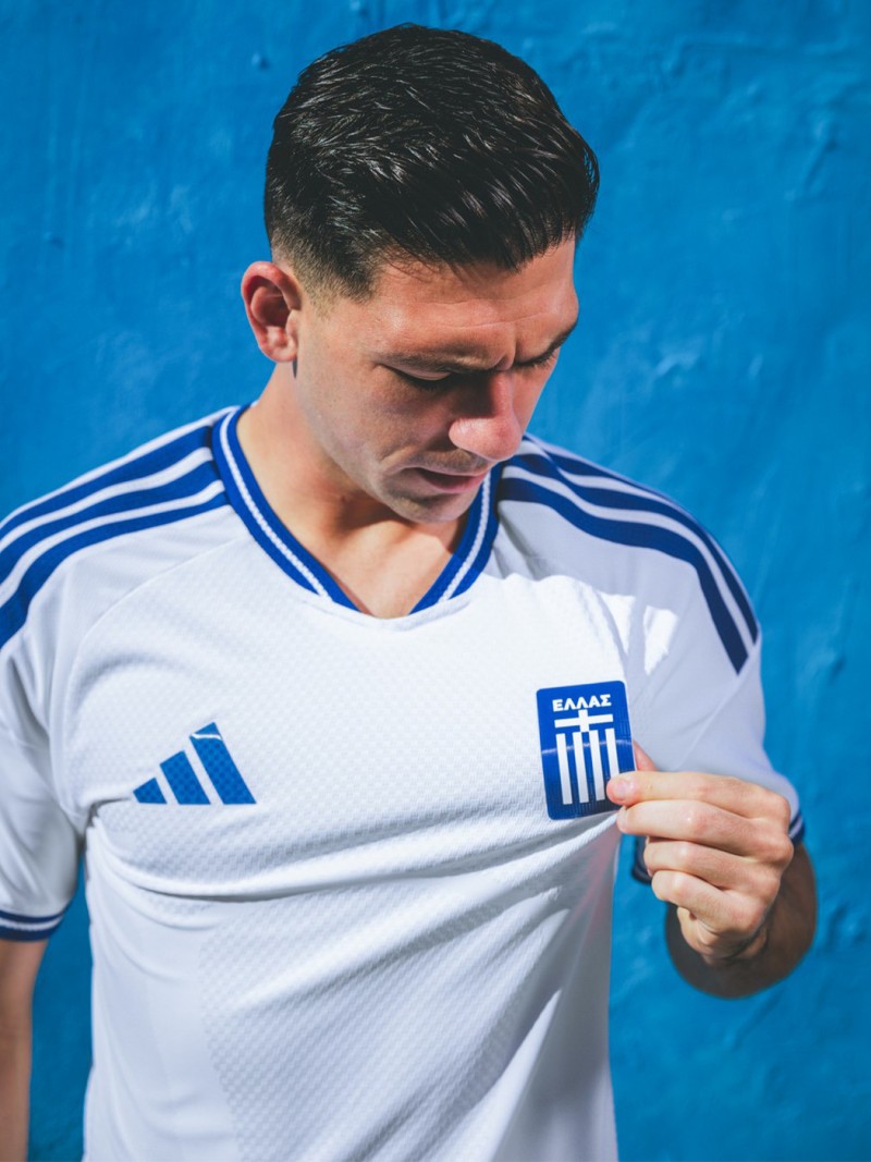 A young man wearing a white and blue soccer jersey stands in front of a vibrant blue background.