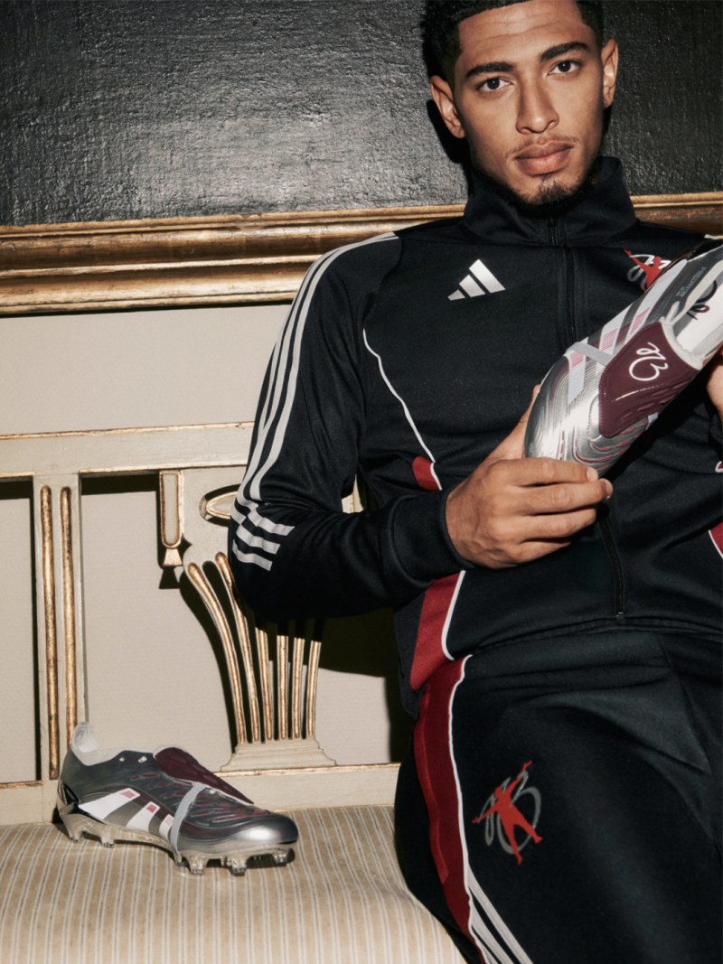 Jude Bellingham wearing a black and red athletic outfit is seated on an ornate, upholstered bench against a backdrop of intricate, patterned carpeting and decorative architectural elements.
