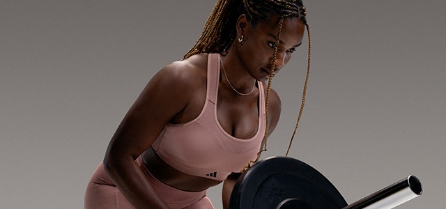 A person with braided hair wearing a pink sports bra and shorts, standing against a plain gray background.