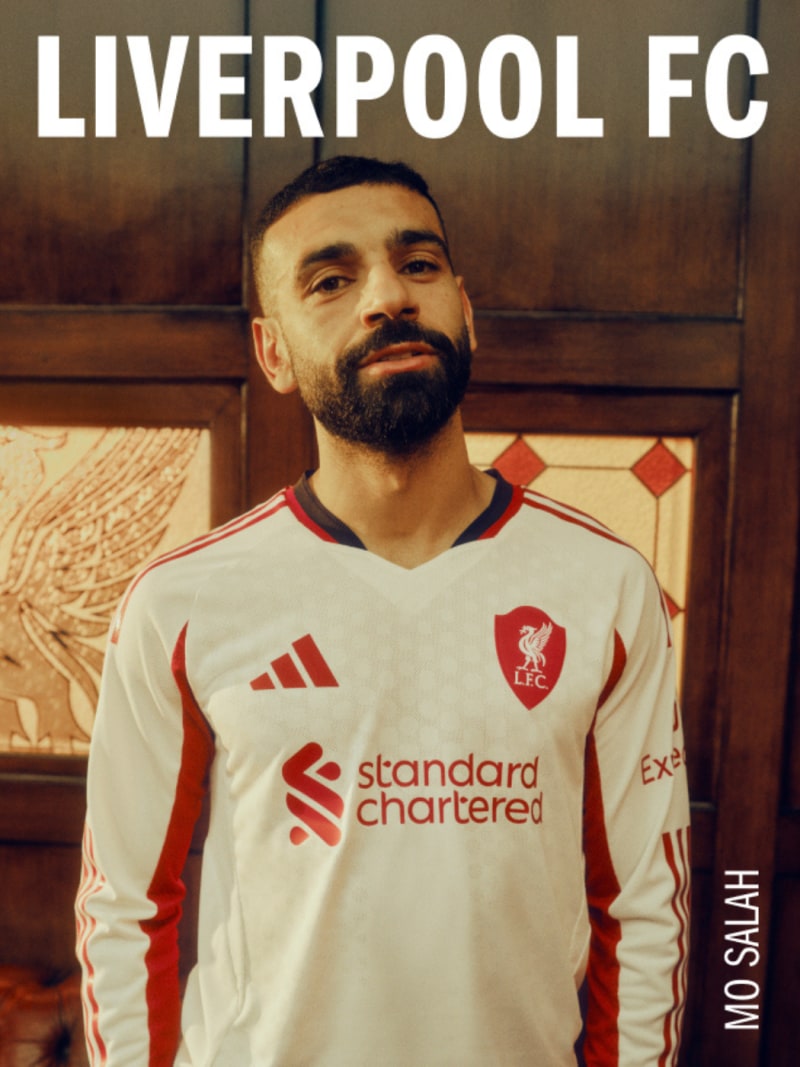The image shows a man wearing a Liverpool FC jersey, standing in front of a wooden background with the Liverpool FC logo prominently displayed.