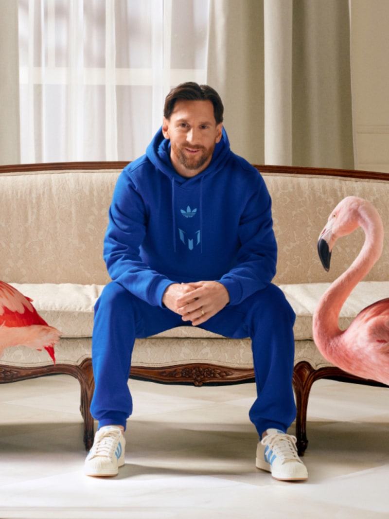 A man in a blue Adidas hoodie and sweatpants sits on a couch, with a large inflatable pink flamingo in the background.
