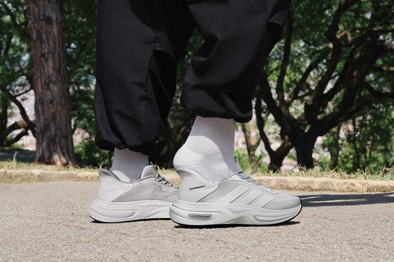 The image shows a person's lower body wearing black pants and gray sneakers, standing on a paved path surrounded by trees in the background.