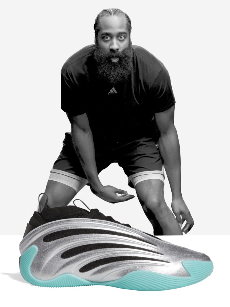 A large, bearded man in a black shirt is crouched down, examining a pair of futuristic-looking sneakers with distinctive wave-like patterns on the soles.