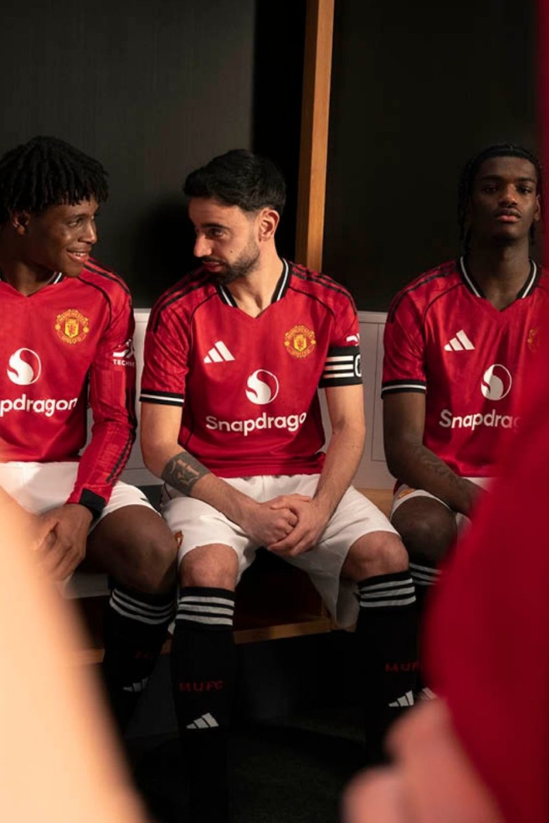 Three individuals wearing Manchester United football jerseys are seated together, with the background appearing to be a locker room or similar indoor setting.