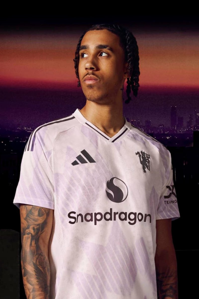 A young man wearing a white soccer jersey with the Snapdragon logo stands in front of a cityscape at sunset.