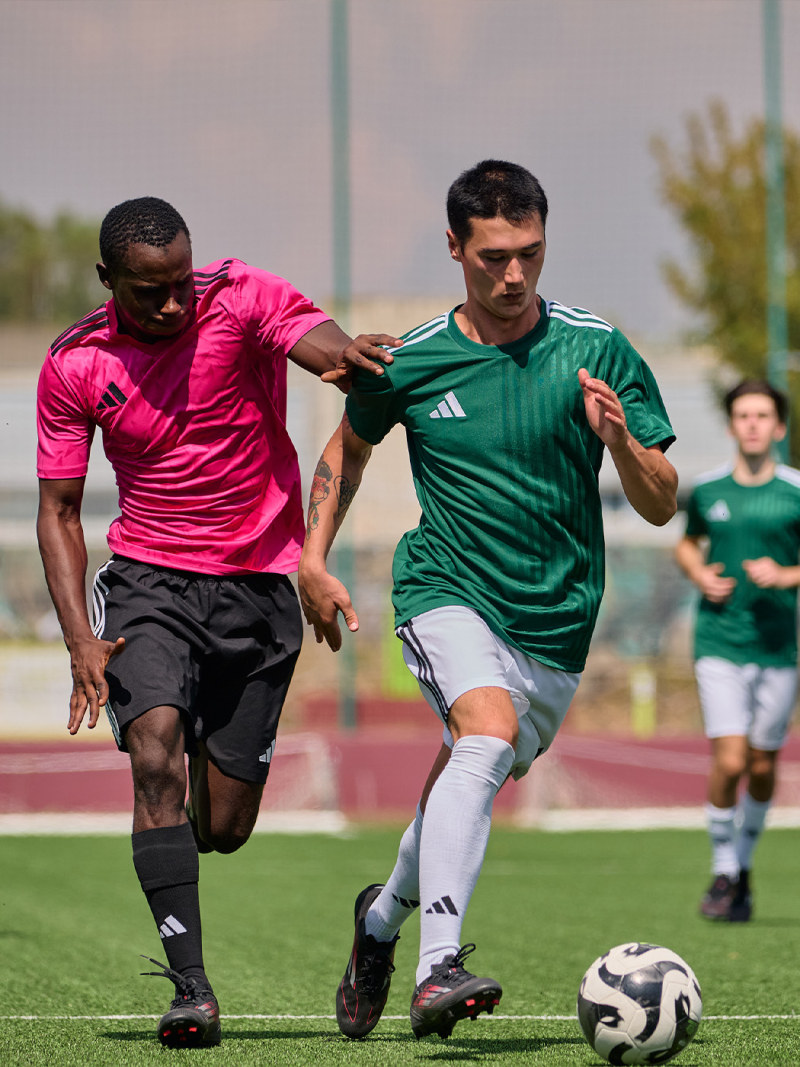 Two soccer players, one in a pink jersey and the other in a green jersey, are engaged in a competitive play on a soccer field with spectators in the background.