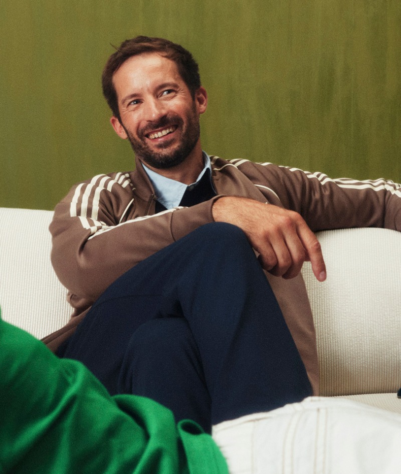 A smiling man with a beard sits comfortably on a couch, wearing a striped shirt and dark pants against a green background.