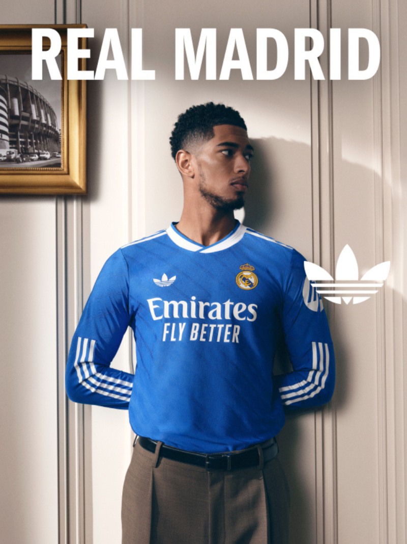 A young man wearing a blue Real Madrid soccer jersey stands in front of a framed image on the wall, with the "Real Madrid" text prominently displayed above him.