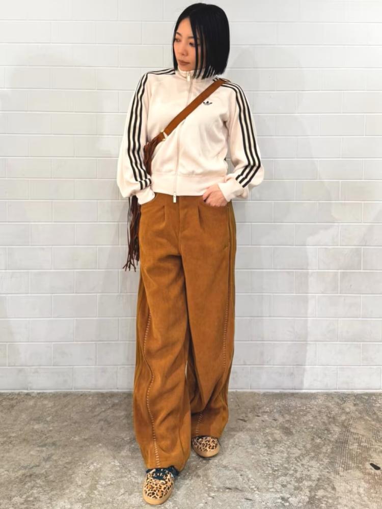 A young woman with short dark hair stands against a plain white wall, wearing a white blouse with black stripes and brown wide-leg pants, accessorized with a brown leather shoulder bag.