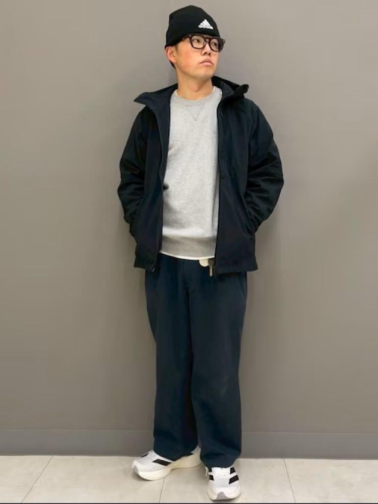 A person wearing a black jacket, white shirt, and black pants stands in a plain, light-colored room.