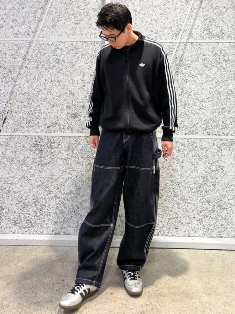 A young person wearing a black Adidas sweatshirt and pants, standing in front of a plain, light-colored wall.