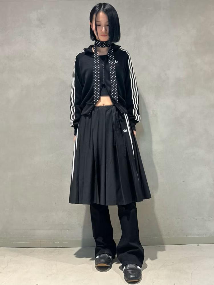 A person wearing a black jacket, crop top, and a long black skirt stands in front of a plain gray background.