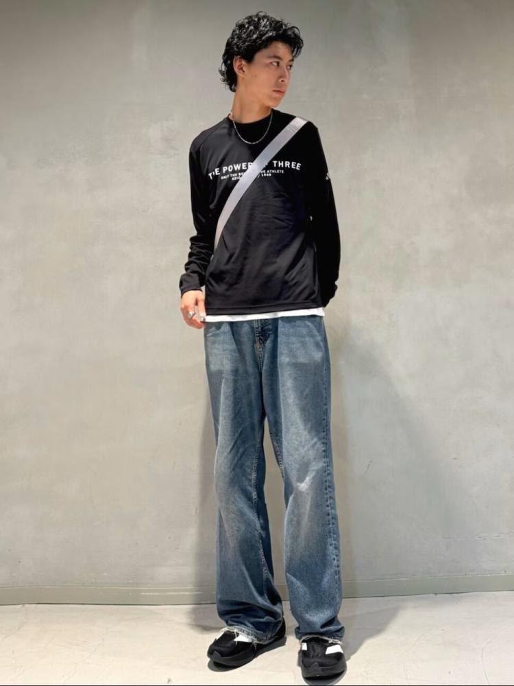 A young person wearing a black sweatshirt and loose-fitting jeans stands against a plain gray background.