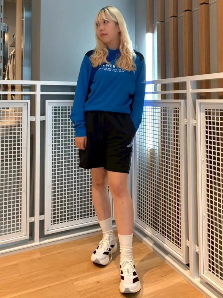 A young woman with blonde hair is standing in a hallway, wearing a blue sweater and black shorts, and holding a pair of white sneakers.